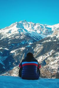 Rear view of boy sitting on snow covered land against mountains and sky