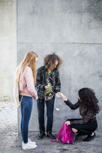 Teenage girl crouching while giving charger to friend against wall