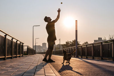 Young man is playing with his dog on empty street in the morning in backlight.