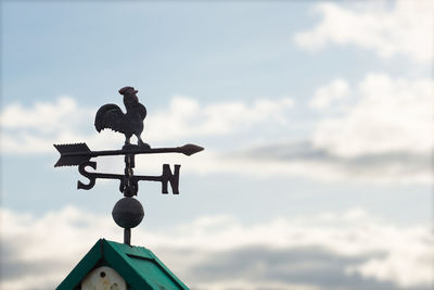 Weather vane on rooftop against cloudy sky