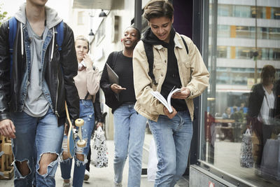 Young man holding book while walking with teenage friends in city