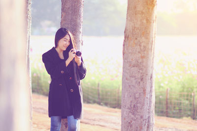 Smiling young woman photographing through camera while standing by tree trunk