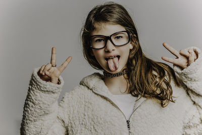 Close-up portrait of cute girl showing peace sign while sticking out tongue against gray background