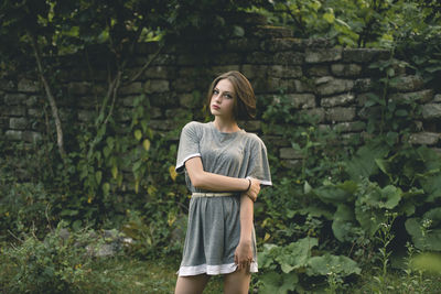 Portrait of young woman standing against plants and wall