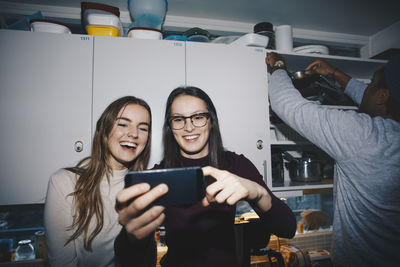 Happy women enjoying with mobile phone by man in kitchen at college dorm
