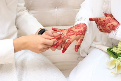 Midsection of groom holding bride finger sitting on sofa during wedding