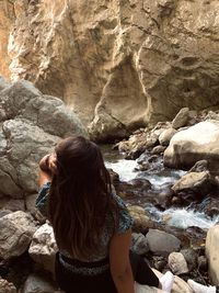 Rear view of girl sitting by flowing water amidst rocks