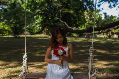 Young woman holding roses sitting on swing at land