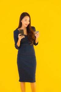 Young woman using phone while standing against yellow background