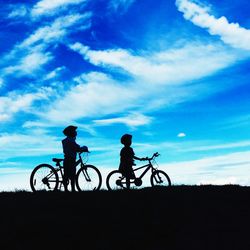 Silhouette children with bicycles standing on field against blue sky