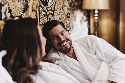 Happy mid adult man talking with girlfriend while relaxing on bed in hotel room