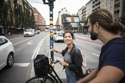 Smiling woman with bicycle looking at friend while standing amidst city street