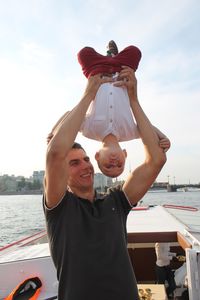 Smiling father carrying son upside down on boat in lake against sky