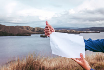 Cropped hands of man holding paper at lake against sky