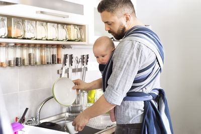 Father with baby son in sling at home doing the dishes