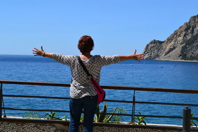 Rear view of woman standing with arms outstretched by railing against sky