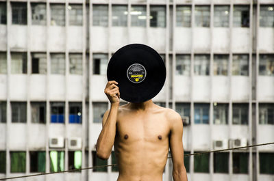 Shirtless man covering his face with record against building