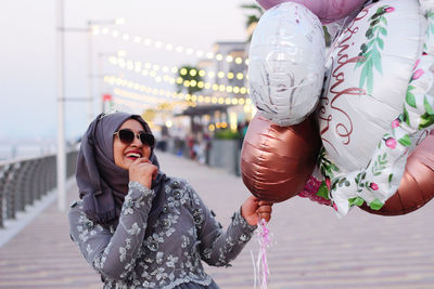 Young woman wearing hijab holding balloons while standing on footpath