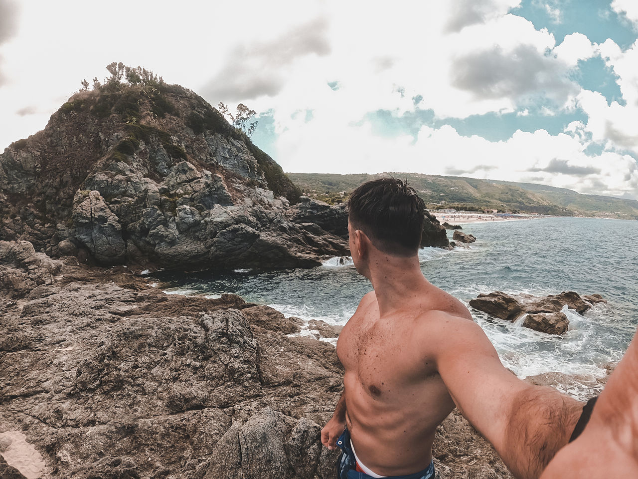 REAR VIEW OF SHIRTLESS MAN ON ROCK AT SHORE