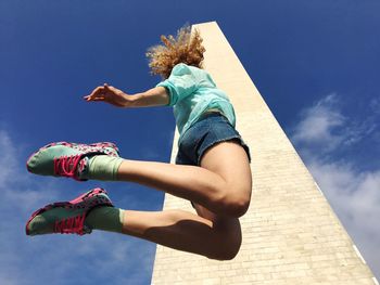 Low angle view of young woman jumping by building against sky