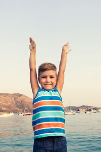 Portrait of smiling boy standing at beach with arms raised against clear sky