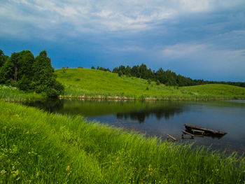 Scenic view of lake by grassy hill against cloudy sky