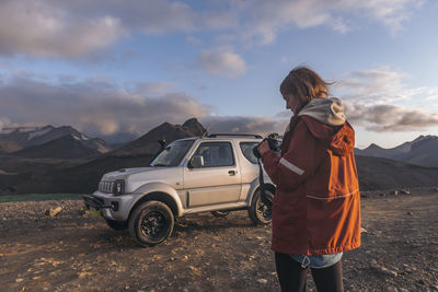 Young woman looking down at camera in front of car in mountains