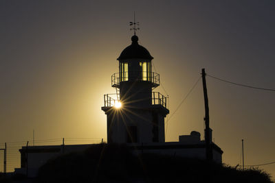 Low angle view of silhouette lighthouse by building against sky during sunset