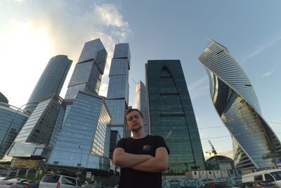 Portrait of young man with arms crossed standing against modern buildings in city