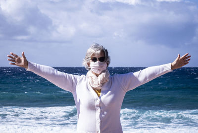 Woman wearing mask with arms outstretched standing by sea against sky