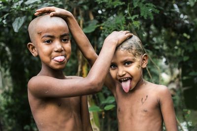 Shirtless siblings with shaved heads sticking out tongues while standing outdoors