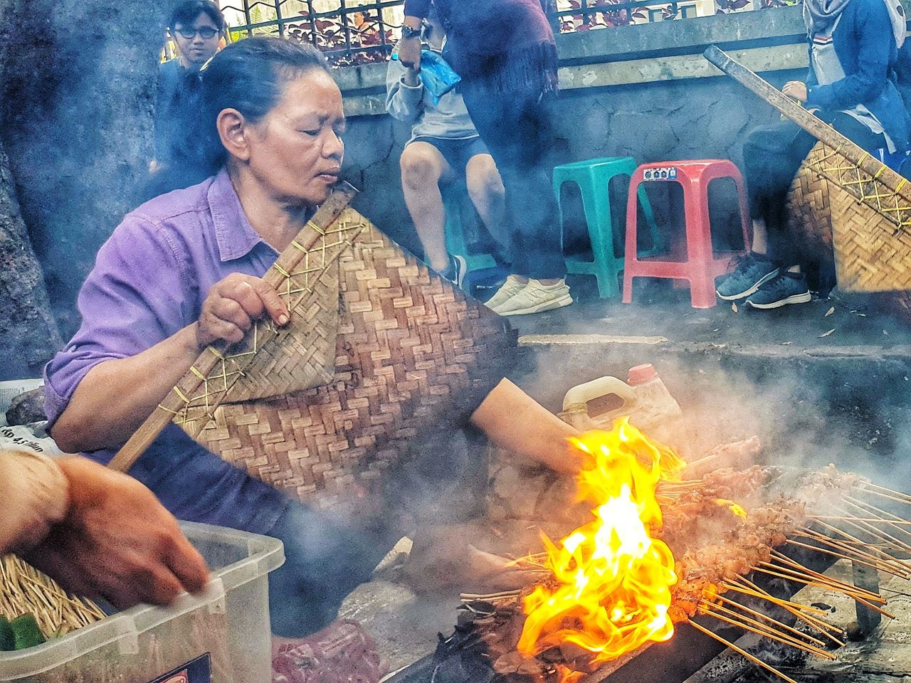 smoke - physical structure, men, real people, people, adult, food, burning, heat - temperature, food and drink, occupation, fire, flame, holding, sitting, day, waist up, preparation, lifestyles, outdoors, preparing food