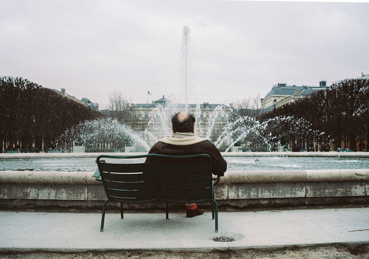 rear view, one person, nature, sky, seat, architecture, sitting, plant, day, tree, winter, real people, bench, water, outdoors, fountain, lifestyles, leisure activity, warm clothing