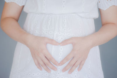 Midsection of pregnant woman making heart shape on stomach while standing against gray background