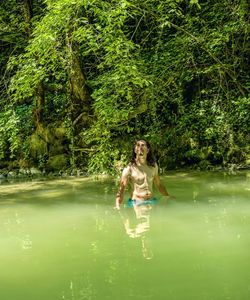 Young man with long black hair, waist-deep in water in a rainforest.