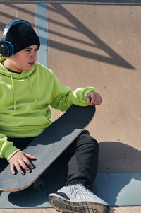Young man, teenager, skateboarding, sitting on the track, wearing headphones, green sweatshirt, 