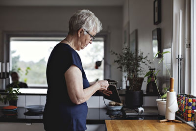 Side view of senior woman charging digital tablet in kitchen at home