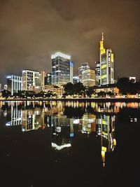 Illuminated buildings by river against sky at night