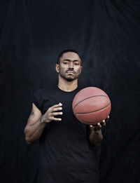 Portrait of man holding basketball while standing against black wall