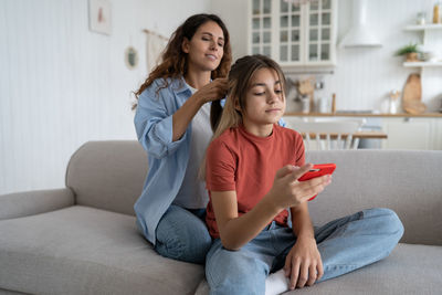 Attentive friendly woman taking care of daughter hairstyle and combing hair sits on sofa at home