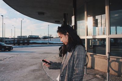 Side view of teenage girl using phone while standing by building