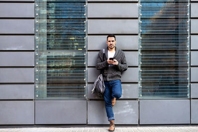 Front view of a bearded man using phone leaning on office building wall