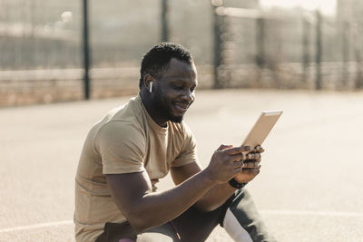 Smiling handsome man using digital tablet while sitting on sports court during sunny day