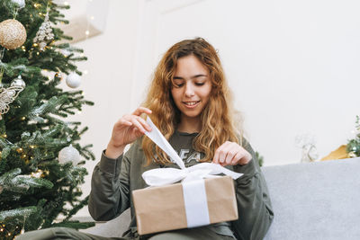 Teenage girl with curly hair holds gift box sitting on sofa in the living room with christmas tree