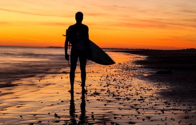 Rear view of silhouette man with surfboard standing on shore at beach during sunset