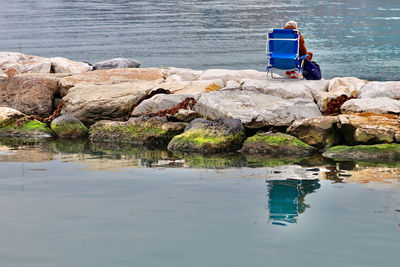 Rear view of woman sitting on chair on rock by sea