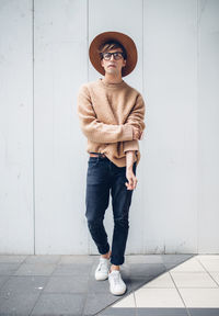 Full length portrait of young man wearing hat while standing against wall