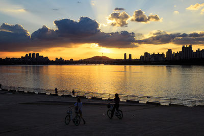 People riding bicycle on river by city against sky during sunset