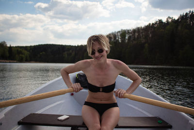 Happy woman wearing black bikini oaring boat in lake against sky