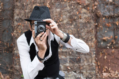 Mature man photographing while standing against brick wall during autumn